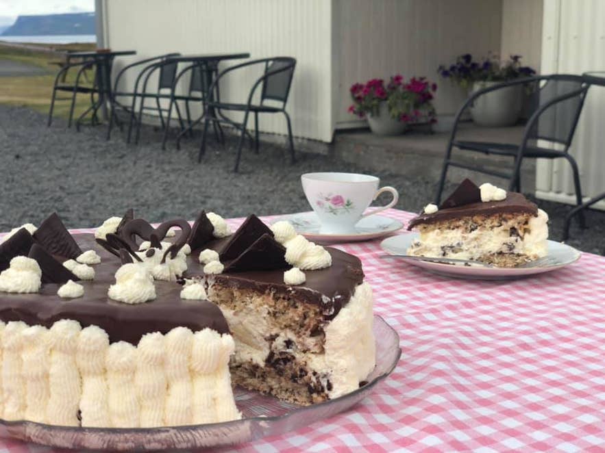 A dessert spread with a whole cake, a slice of cake on a separate plate, and a teacup, set outdoors at Ogur Cafe in Sudavik