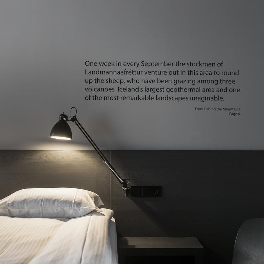 A lamp shines over a comfortable bed, with a book excerpt on the wall at Skuggi Hotel Reykjavík by Keahotels. 