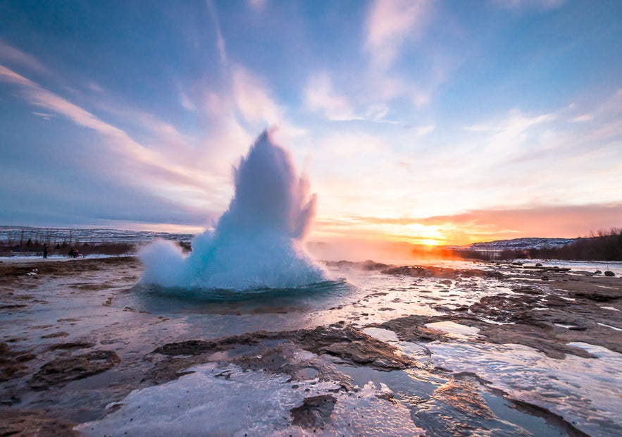 Strokkur geyser erupting at sunrise in the Golden Circle, Iceland, with steam rising and colorful skies reflecting on frozen ground.