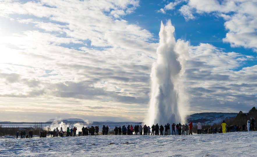 A crowd watches Strokkur geyser erupt against a bright winter sky in the Haukadalur Geothermal Area.