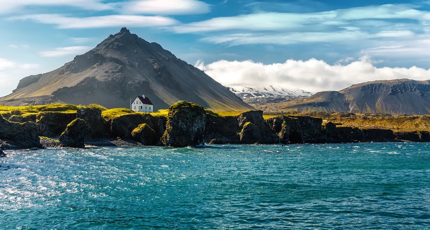 White house near basalt rocks and ocean at Arnarstapi with snowy mountains behind, close to the Londrangar cliffs.