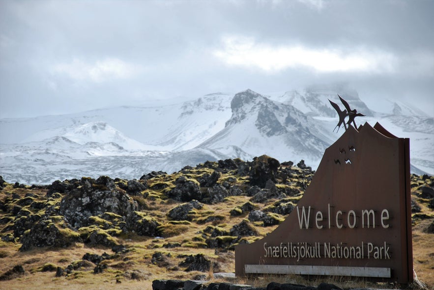 Welcome sign at Snæfellsjökull National Park with mossy lava rocks in foreground and snow-covered mountains in background.