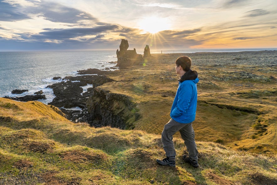 "Man in blue jacket standing on grassy edge of Londrangar cliffs at sunset, overlooking sea stacks and rugged coastline