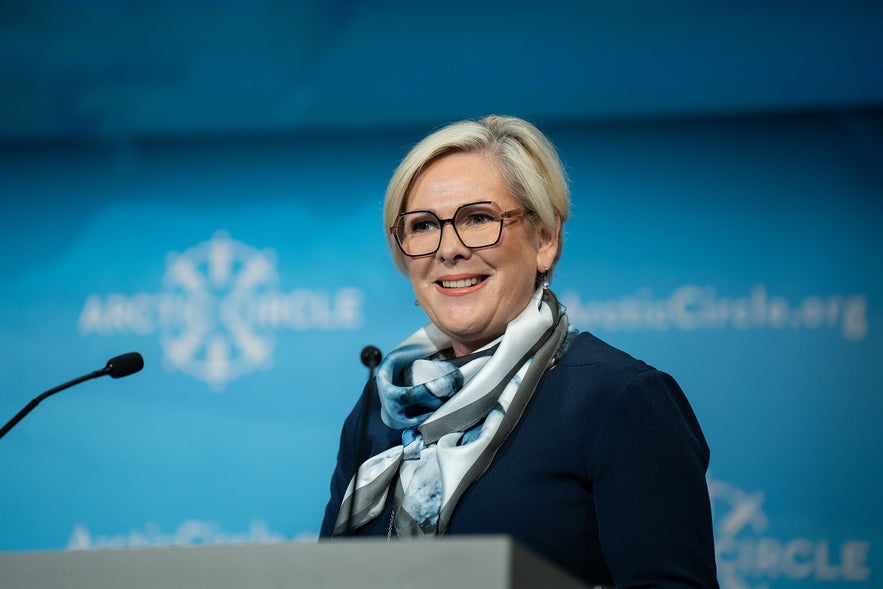 Halla Tómasdóttir speaking at the Arctic Circle conference, wearing glasses and a patterned scarf, standing at a podium with a blue background and Arctic Circle logo.