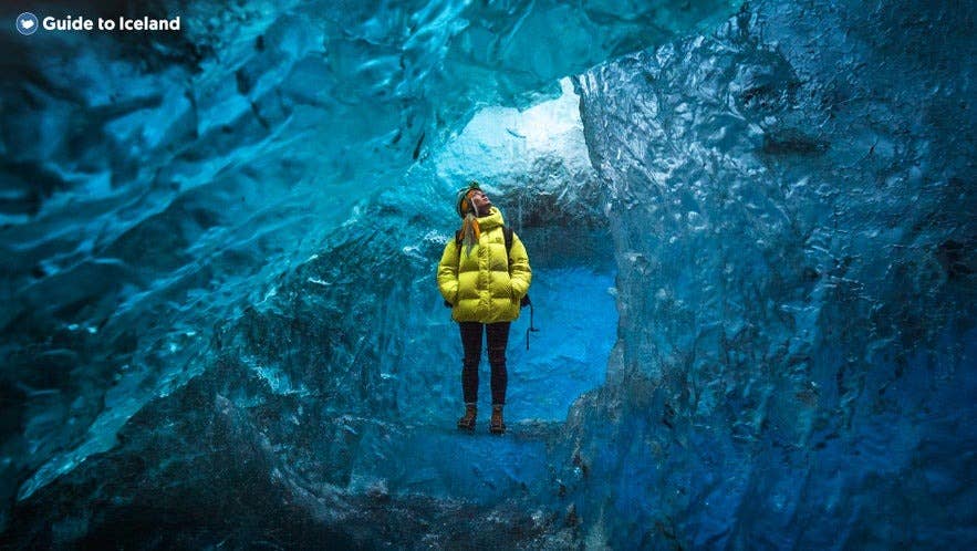 Reiziger in gele jas verkent een kristalblauwe ijsgrot bij de Jokulsarlon-gletsjerlagune in IJsland, omringd door felblauwe gletsjerwanden en natuurlijk licht dat door het ijs schijnt.