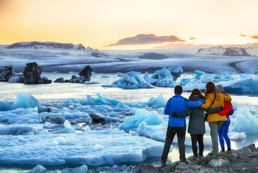 Group of friends standing arm-in-arm, admiring floating icebergs at Jokulsarlon Glacier Lagoon in Iceland during a colorful winter sunset, with snow-covered mountains and glacial landscapes in the background.