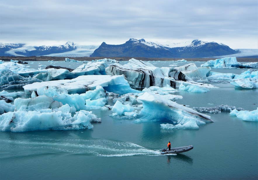 Kleine opblaasboot met een persoon in oranje jas die tussen drijvende blauwe ijsbergen vaart op de Jokulsarlon-gletsjerlagune in IJsland, met besneeuwde bergen en gletsjers op de achtergrond onder een bewolkte lucht.