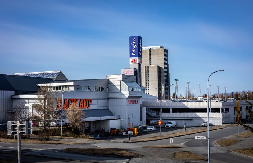 Exterior view of Kringlan Shopping Mall in Reykjavik, Iceland, showing major retail stores like H&M, Hagkaup, and Lindex, with modern architecture and parking facilities under a clear blue sky.