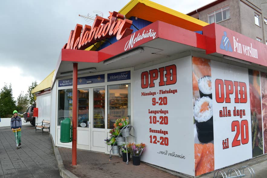 Außenansicht des Supermarkts Melabudin in Reykjavik, Island, mit auffälligem roten Schild, Öffnungszeiten am Eingang, Blumenstand und einem Kind auf dem Roller auf dem Gehweg.