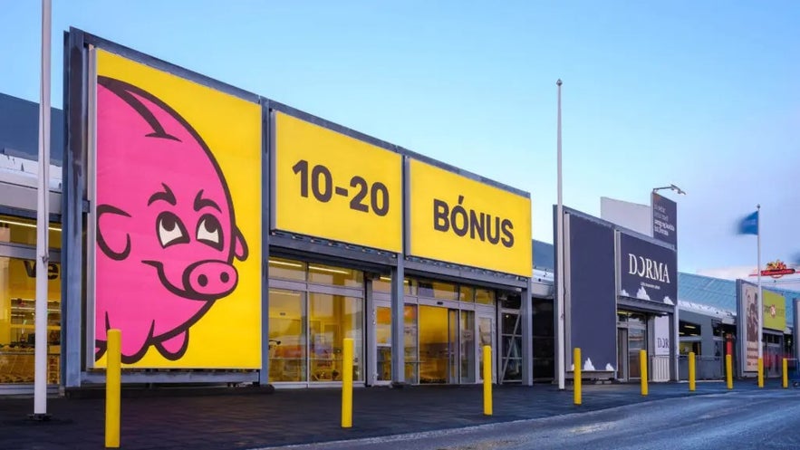 Exterior of a Bónus grocery store in Iceland featuring the signature bright yellow signage with a smiling pink piggy bank logo and store hours displayed as 10–20.
