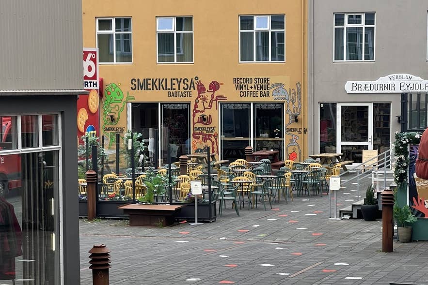Udsigt til pladebutikken og kaffebaren Smekkleysa ("dårlig smag") i Reykjavik, Island, med markant gadekunst, farverige siddepladser på terrassen og skilte, der reklamerer for vinylplader, live-musik og kaffe i gårdhaven Hjartatorg.