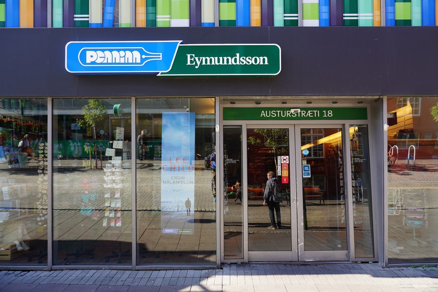 Front entrance of Penninn Eymundsson bookstore on Austurstræti Street in downtown Reykjavik, Iceland, with glass windows displaying books and colorful architectural panels above.