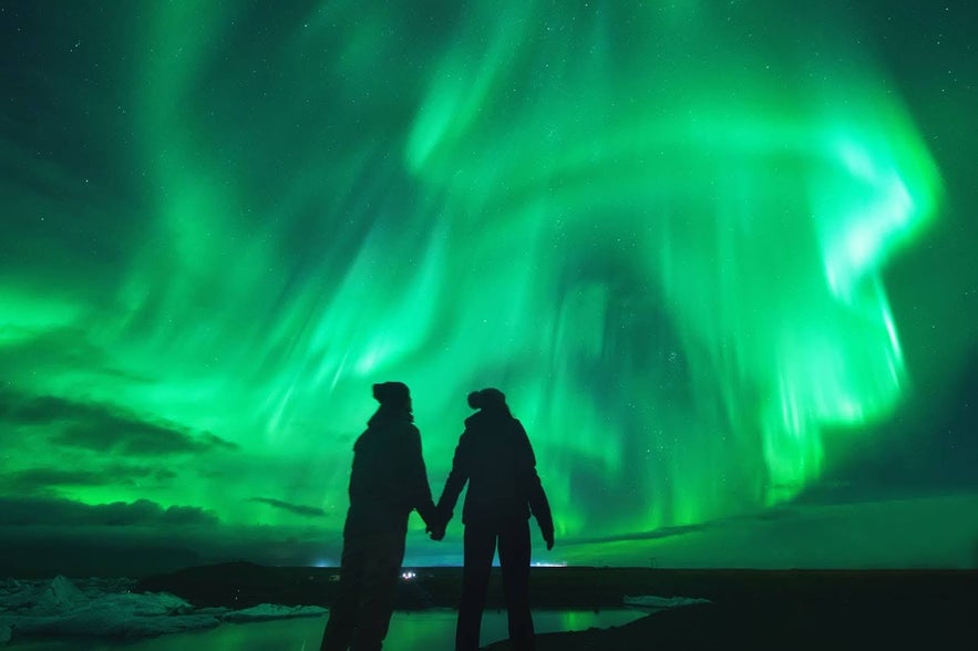 Couple holding hands while watching the northern lights in Iceland, with vivid green auroras dancing across the night sky.