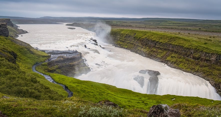 Gullfoss Waterfall in Iceland with mist rising from the powerful cascade and visitors walking along a green path leading to the viewpoint.