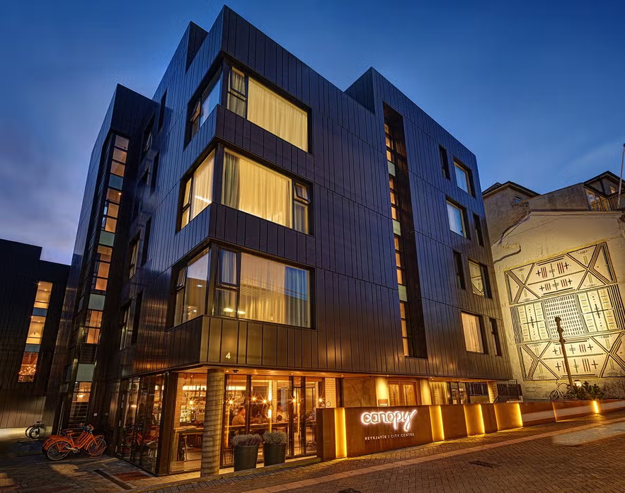 Exterior of Canopy by Hilton Reykjavik with warm lighting, modern design, and a couple walking toward the entrance on a quiet evening.