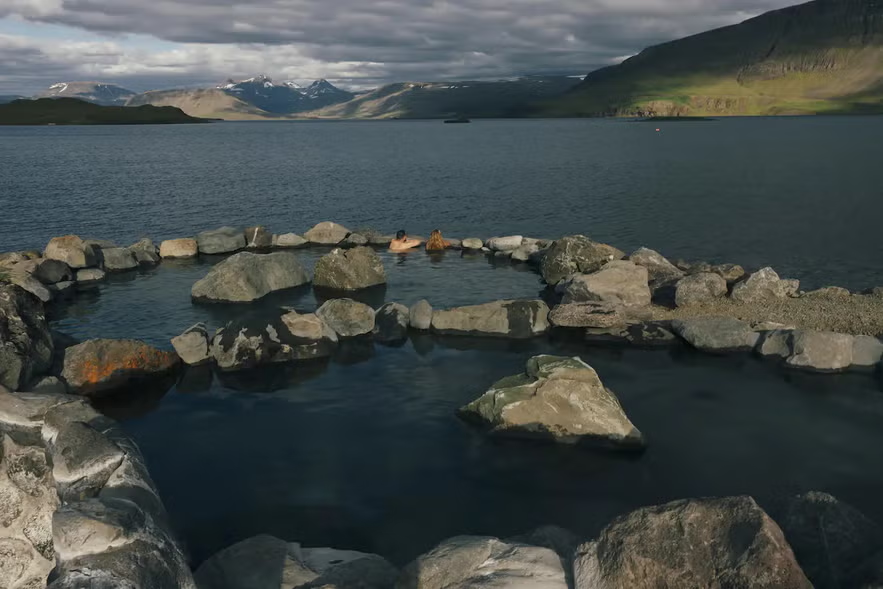 Two people relaxing in a rock-lined hot spring at Hvammsvík, Iceland, with views of the fjord and distant mountains.