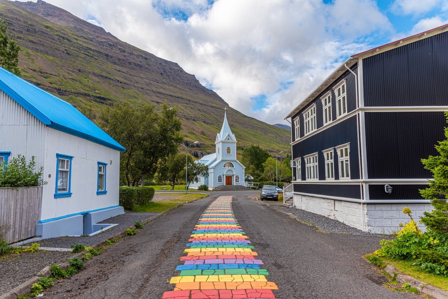 Rainbow-painted street leading to a blue church in Seydisfjordur, East Iceland, with mountains in the background.