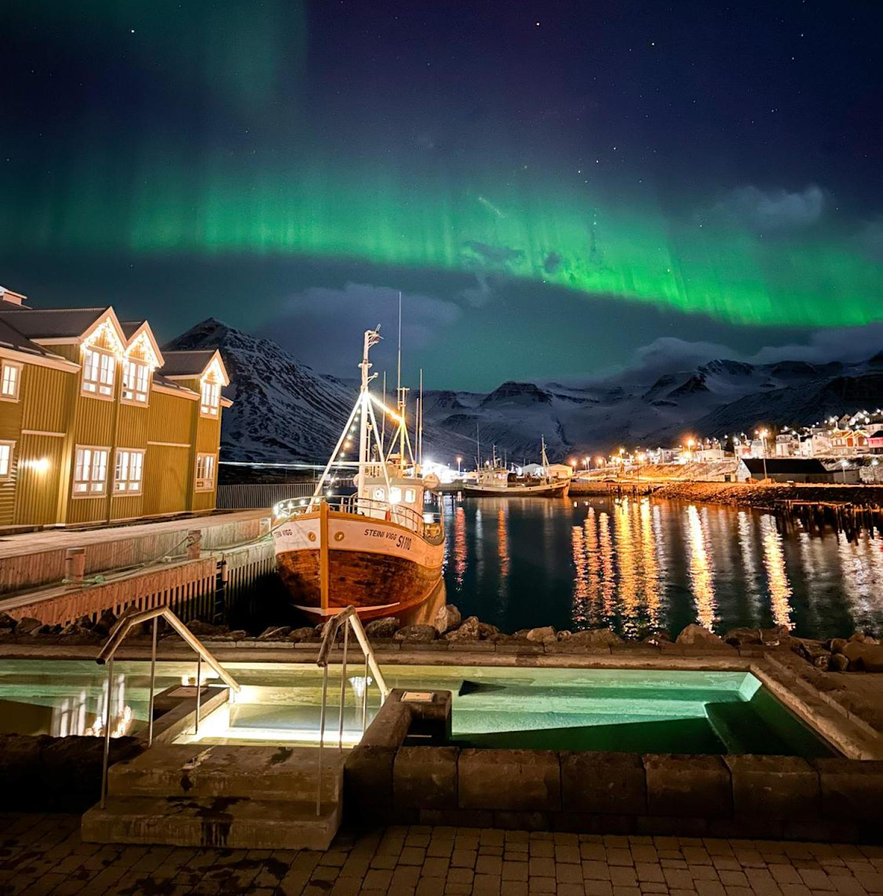 Northern lights above Siglo Hotel in Siglufjordur, Iceland, with fishing boats docked beside the yellow waterfront building and outdoor hot tubs overlooking the snowy mountains and harbor.