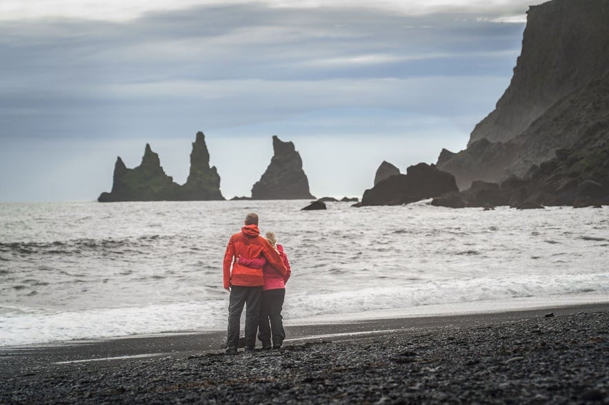 Couple in red jackets standing on Reynisfjara black sand beach in Iceland, facing the Reynisdrangar sea stacks.