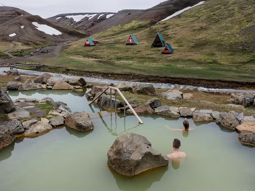 Two people bathing in a natural geothermal pool, with colorful A-frame cabins and snow-dusted highlands in the background.