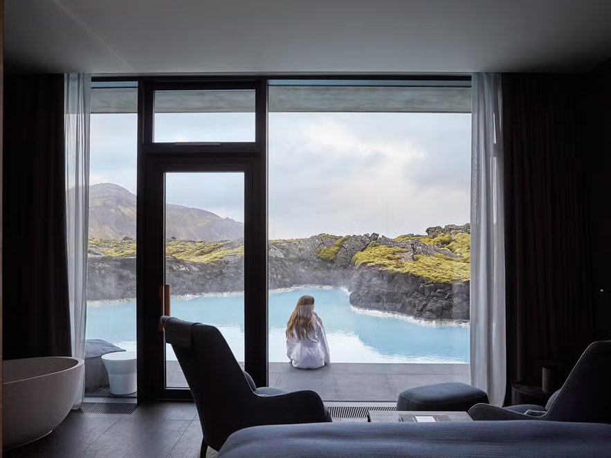 Woman in a robe sitting by the geothermal waters outside a suite at The Retreat at Blue Lagoon, Iceland, with moss-covered lava fields in the background.