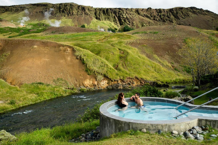 Couple relaxing in an outdoor geothermal hot tub at Frost and Fire Hotel in Hveragerdi, with a scenic river and mossy hills in the background.