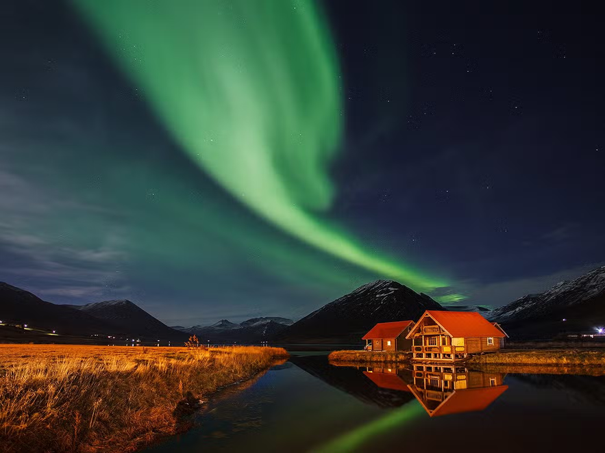 Brimmes Cabins in Iceland with red rooftops reflected in a still lake, set beneath the northern lights and surrounded by mountains.