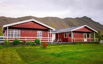 Front view of Moar Guesthouse with red timber siding, picnic table, and a scenic mountain backdrop in West Iceland.
