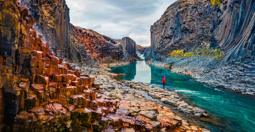 Person standing beside a turquoise glacial river surrounded by towering basalt columns in Studlagil Canyon, East Iceland. Person standing beside a turquoise glacial river surrounded by towering basalt columns in Studlagil Canyon, East Iceland.