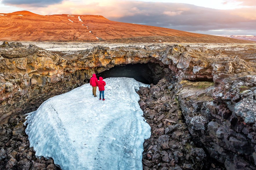 Two people in red jackets walking on ice toward the entrance of Surtshellir lava cave in West Iceland.