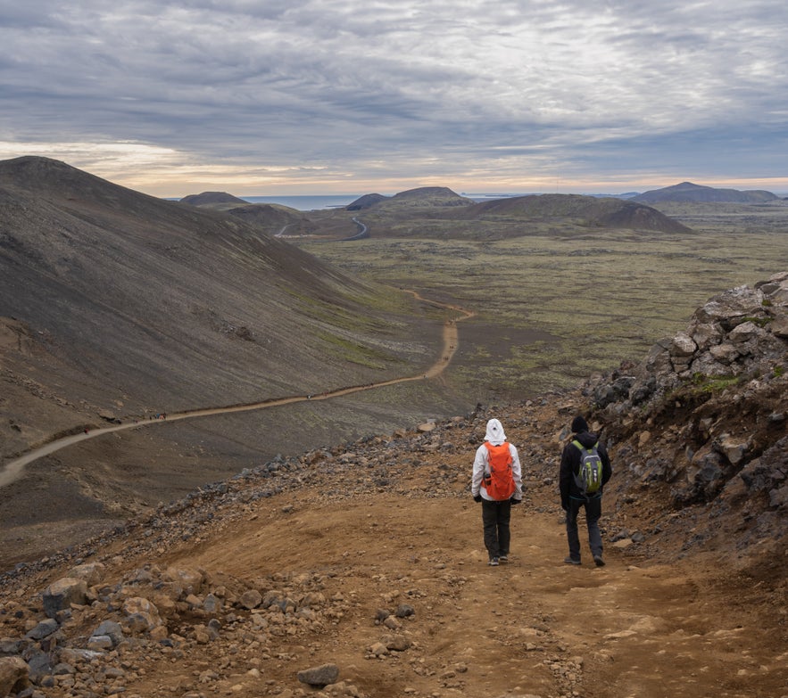 Two hikers descend a rocky trail surrounded by volcanic terrain in the Reykjanes Peninsula, Iceland.