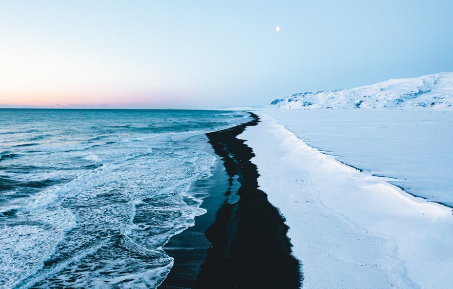 Snow-covered Dyrholaey coastline in Iceland with black sand beach stretching along the Atlantic Ocean under a pale winter sky and a visible moon.