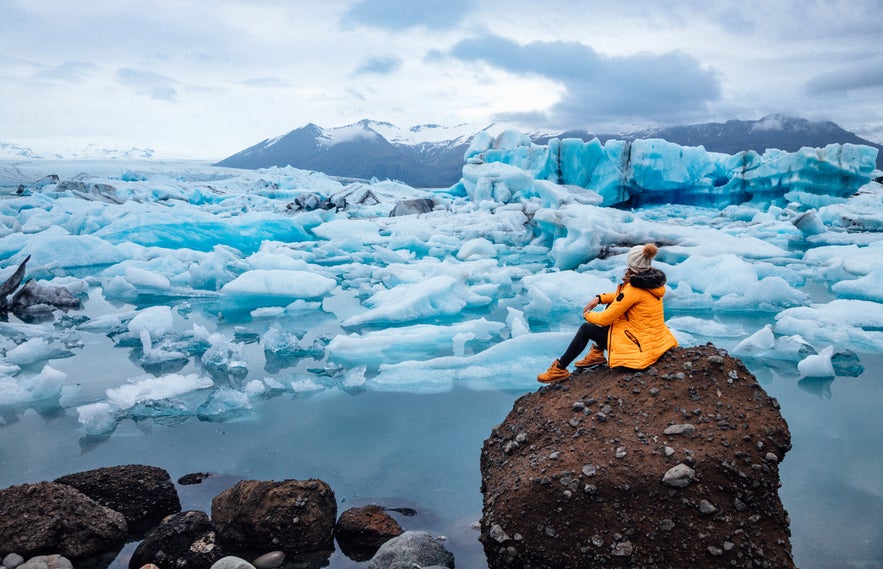 Traveler in yellow jacket sitting by Jokulsarlon glacier lagoon surrounded by floating icebergs in South Iceland.