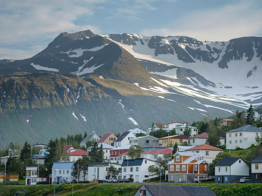 Colorful houses on a hillside in Siglufjörður, Iceland, with dramatic snow-streaked mountains in the background under a partly cloudy sky.