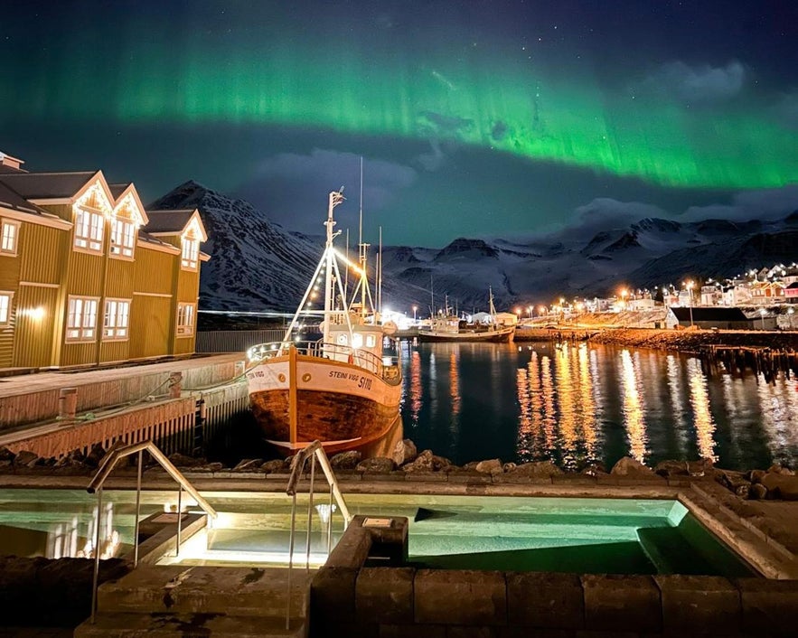 Geothermal hot tub at Siglo Hotel in Siglufjörður, Iceland, overlooking a calm harbor with a wooden fishing boat, under vibrant northern lights, perfect for a peaceful and scenic retreat.