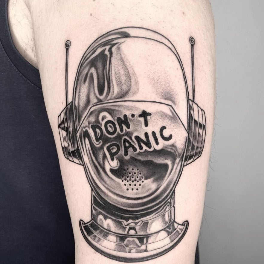 Black and grey tattoo of an astronaut helmet on an upper arm, with the words "DON'T PANIC" written on the visor.