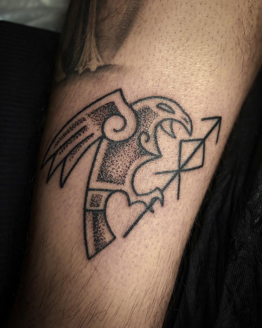 Black ink tattoo of a stylized eagle with geometric shapes, heart, and arrow.