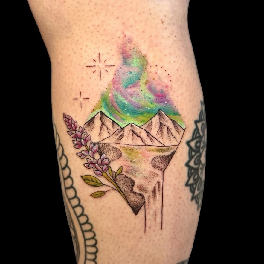 Leg tattoo featuring Icelandic mountains with blooming lupine flowers, a cascading waterfall, and northern lights in the sky.