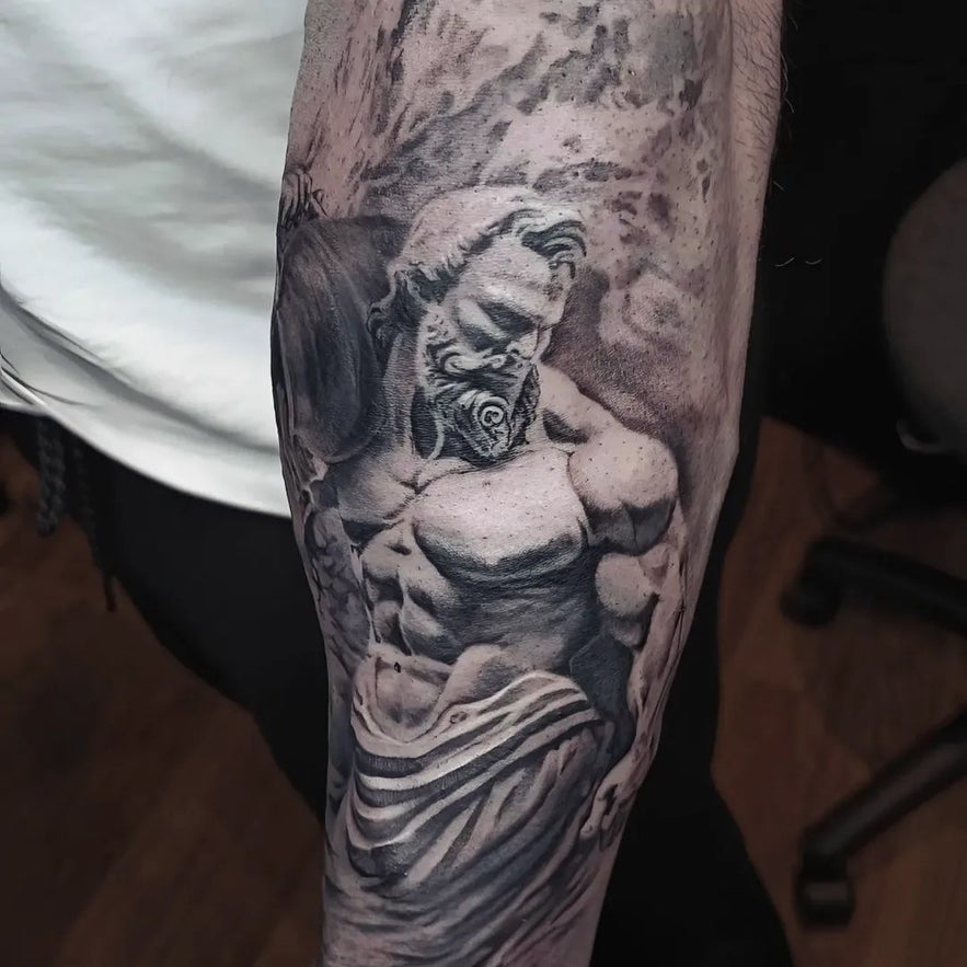 Realistic black and grey arm tattoo depicting a muscular, bearded Greek god figure in classical drapery, with detailed shading and dramatic contrast.