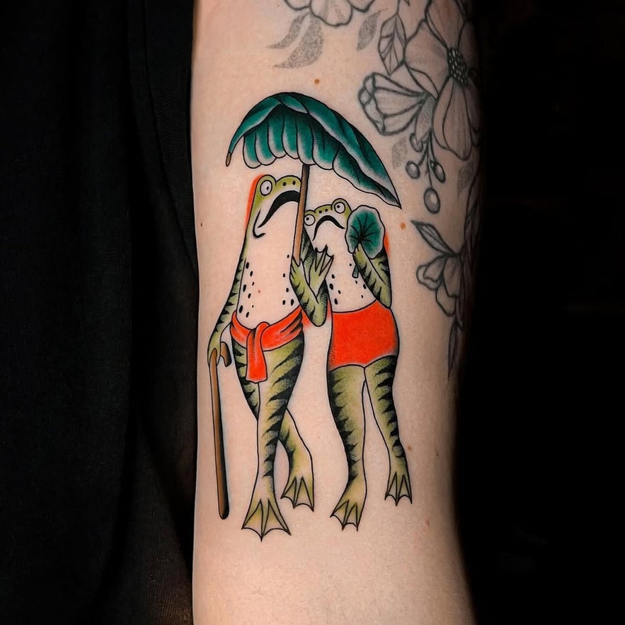 Color tattoo of two green frogs standing, one holding a leaf umbrella and the other holding a leaf fan.