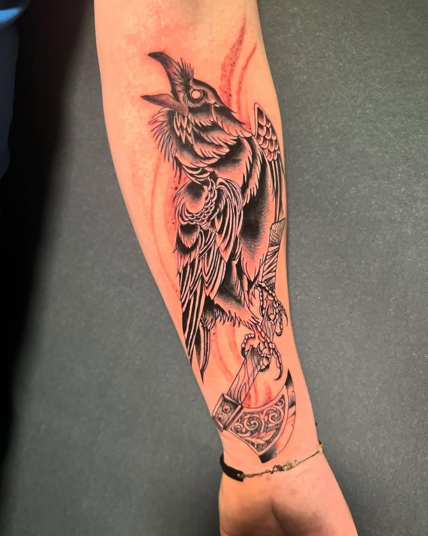 Black and grey forearm tattoo of a stylized raven holding an axe in its talons, featuring bold lines and shading with mythological undertones. Black and grey forearm tattoo of a stylized raven holding an axe in its talons, featuring bold lines and shading with mythological undertones.