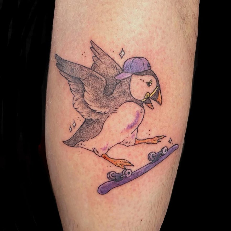 Tattoo of a cartoon-style penguin wearing a cap, doing a kickflip on a skateboard.