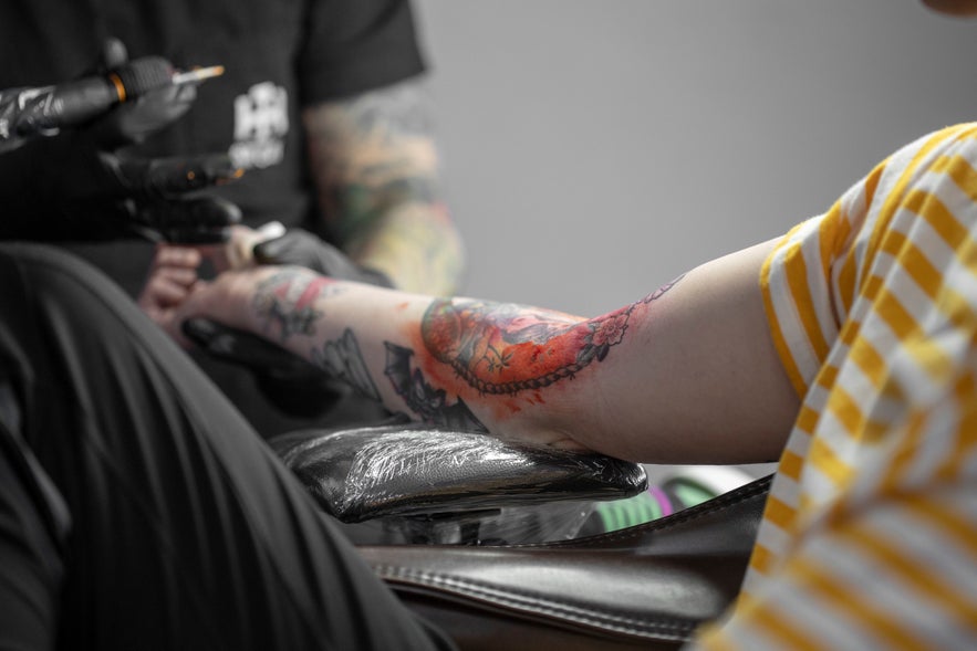 A man with an arm tattoo sits while a tattoo artist adds new ink to the same arm.