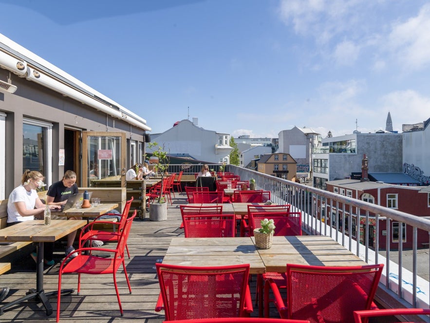 Loft Hostel rooftop bar in Reykjavik on a sunny day, with red chairs and wooden tables lined along the terrace, and guests enjoying drinks or working on laptops with a view of the city.