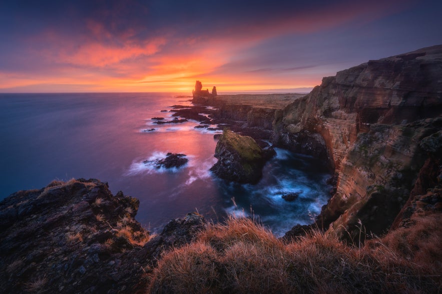 The Londrangar Cliffs and sea stacks in West Iceland are lit by sunset, with crashing waves and dramatic skies.