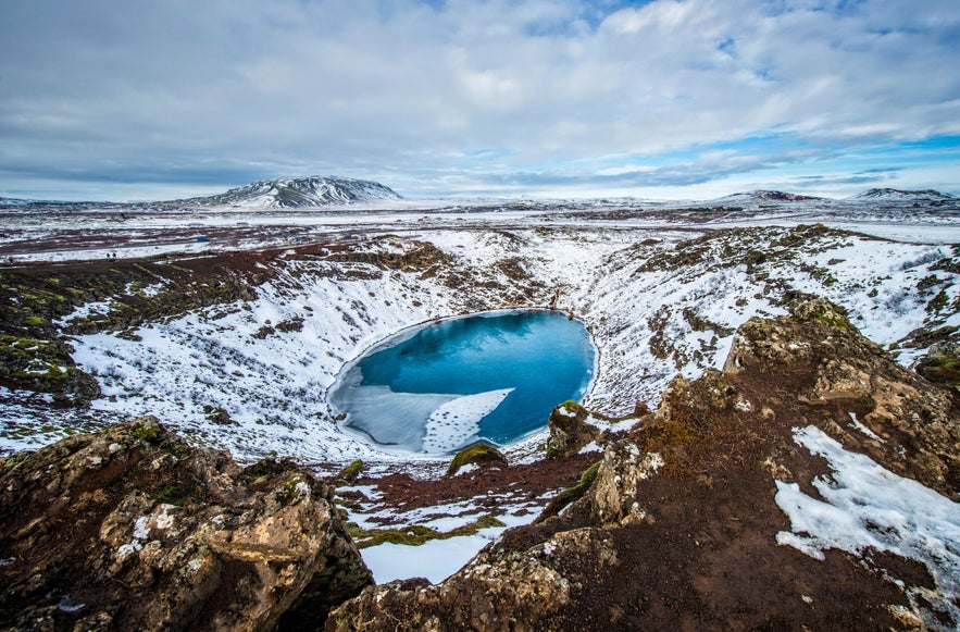 Kerid Crater in winter, with icy blue water and snow-covered slopes contrasting against dark volcanic rock.