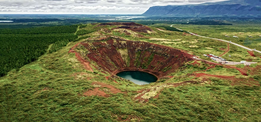 Aerial view of Kerid Crater surrounded by green moss and red earth, with walking trails and a nearby parking lot.