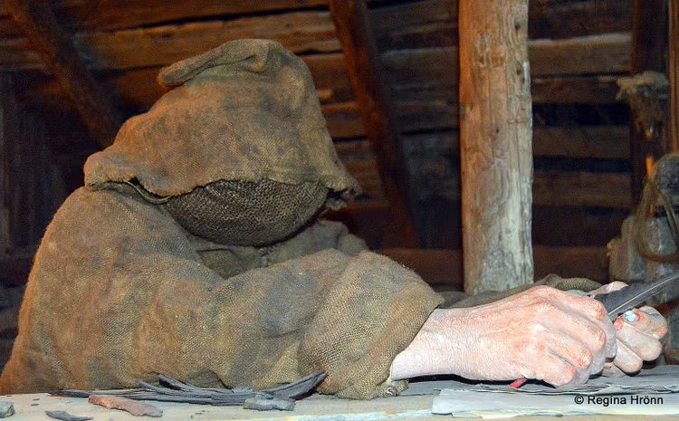 Hooded figure in rough wool cloak, carving magical staves, displayed at the Museum of Icelandic Sorcery and Witchcraft.