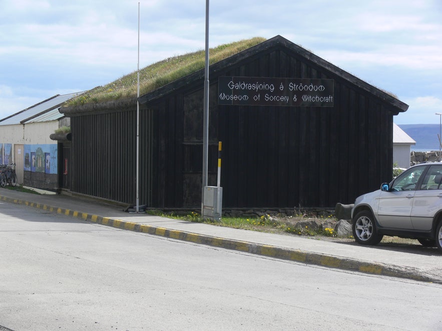Exterior side view of the Museum of Icelandic Sorcery and Witchcraft in Holmavik, Iceland, featuring a turf-covered roof, dark wooden facade, and a sign displaying the museum's name.