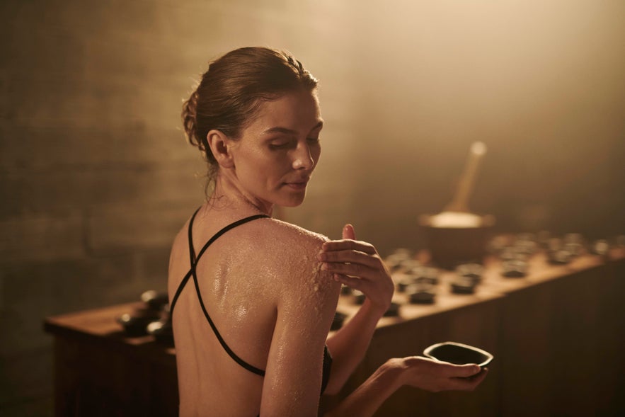 A person with hair tied back applies a body scrub to their shoulder at Sky Lagoon, holding a small bowl. A person with hair tied back applies a body scrub to their shoulder at Sky Lagoon, holding a small bowl.